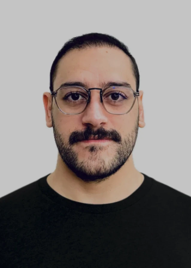 Retrato de Enrico Gastaldelli: homem de óculos de armação transparente, bigode e barba, com camiseta preta, sobre fundo cinza claro.