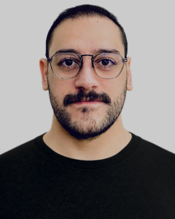 Retrato de Enrico Gastaldelli: homem de óculos, bigode e barba, com camiseta preta, sobre fundo cinza.